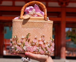 Four Seasons Collection Spring Blossom handcrafted raffia bag with delicate cherry blossom embroidery displayed in front of a traditional Japanese temple.